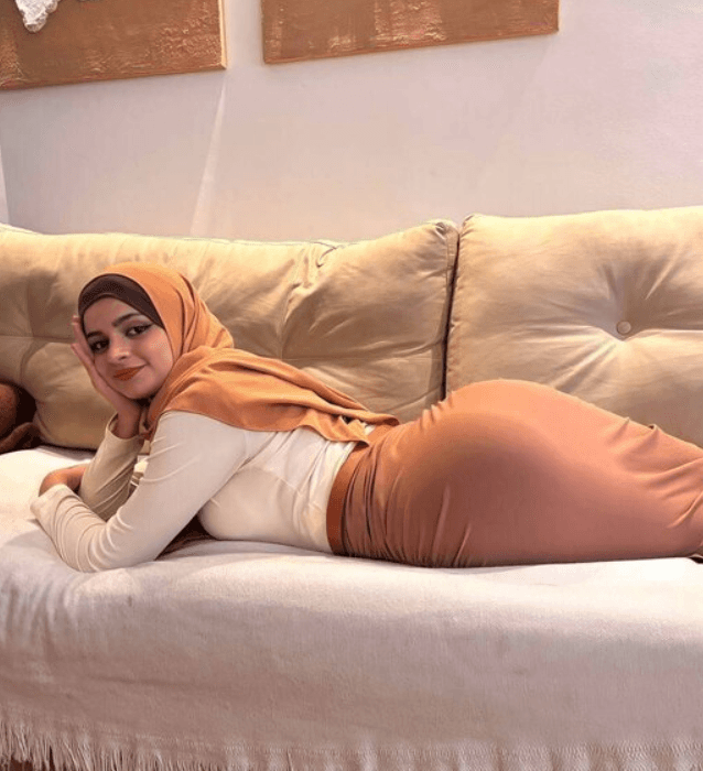 Woman wearing a peach hijab and white top lying on her stomach on a beige couch, resting her head on her hands and looking at the camera.