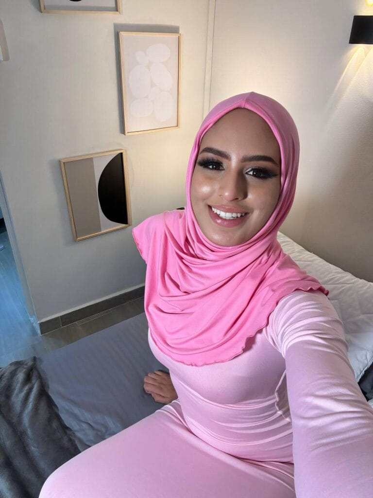 Smiling woman in a pink hijab and matching outfit taking a selfie indoors, with abstract framed art on a light wall behind her.