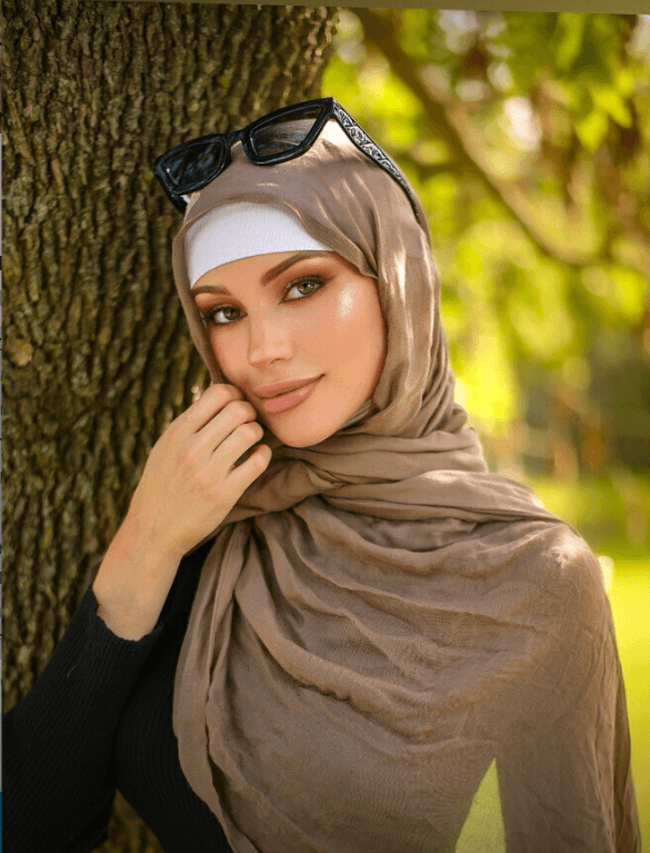 Portrait of a woman in a beige hijab with sunglasses on her head, leaning against a tree in a sunlit park.