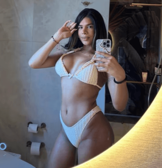 Woman in a white bikini taking a mirror selfie in a bathroom, holding her phone to capture her pose.
