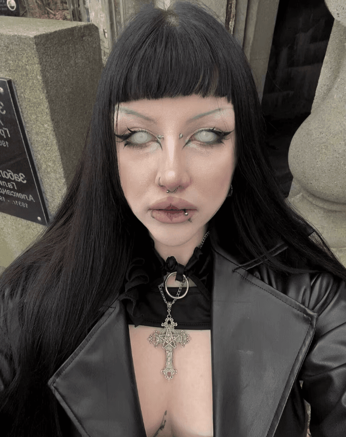 Person with long black hair and blunt bangs wearing a black leather jacket, heavy gothic makeup, facial piercings, and a large cross necklace.