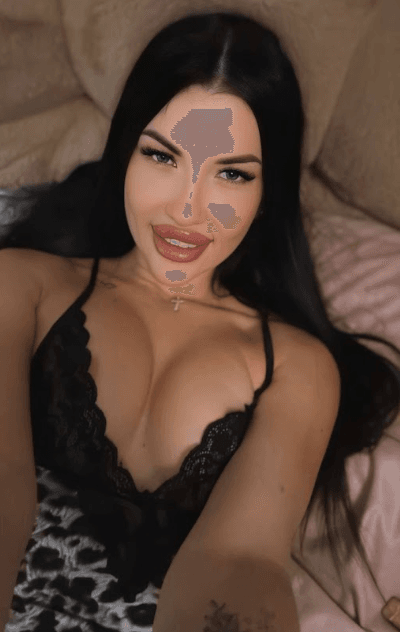 Selfie of a woman lying on a bed, wearing black lace lingerie, looking at the camera with a smile, cross necklace visible.