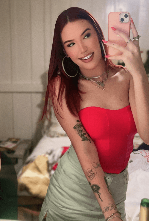 Woman taking a mirror selfie in a red strapless top, showing tattoos on her arm and holding a pink smartphone.