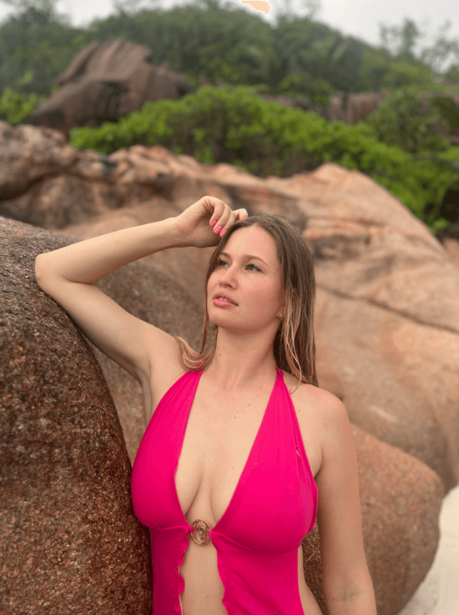 Woman in a hot pink halter swimsuit leaning against a rocky beach wall, gazing into the distance.