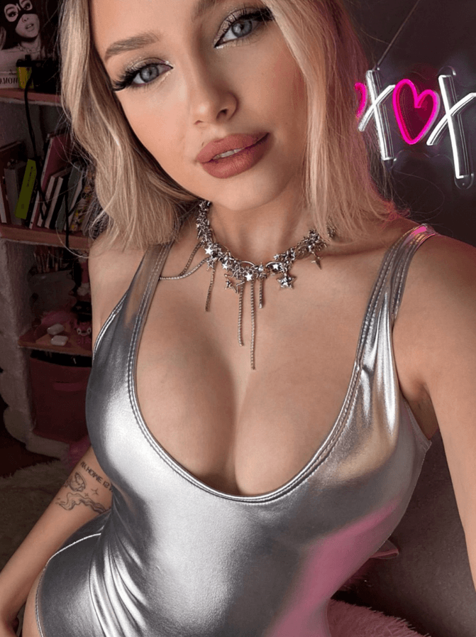 Selfie of a young woman with blonde hair wearing a metallic silver V‑neck top and a star necklace, tattoo on her left arm, neon sign in the background.