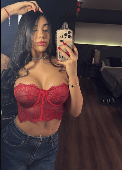 Woman taking a mirror selfie in a red lace bustier and jeans, holding a smartphone with a pink case in front of her.