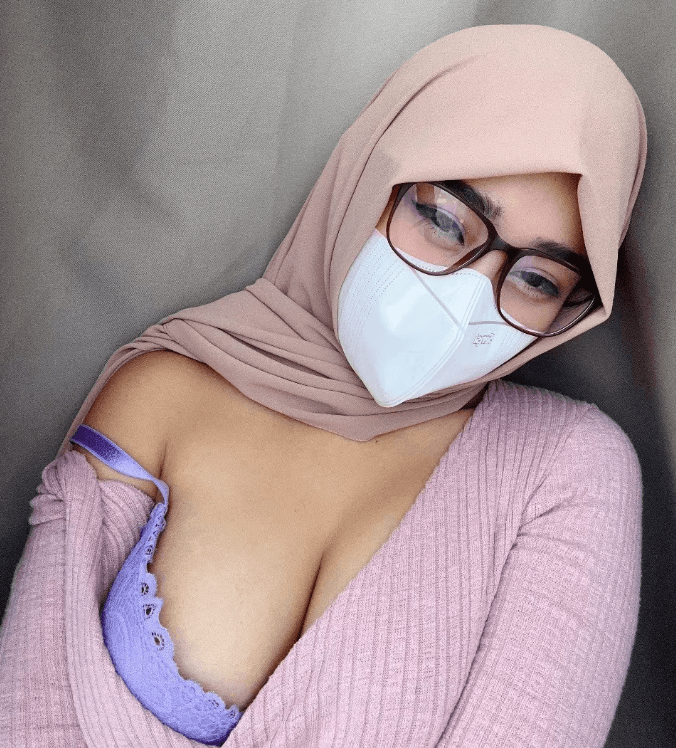 Person wearing a beige hijab, face mask, and glasses, taking a selfie in a light pink sweater; purple bra slightly visible.