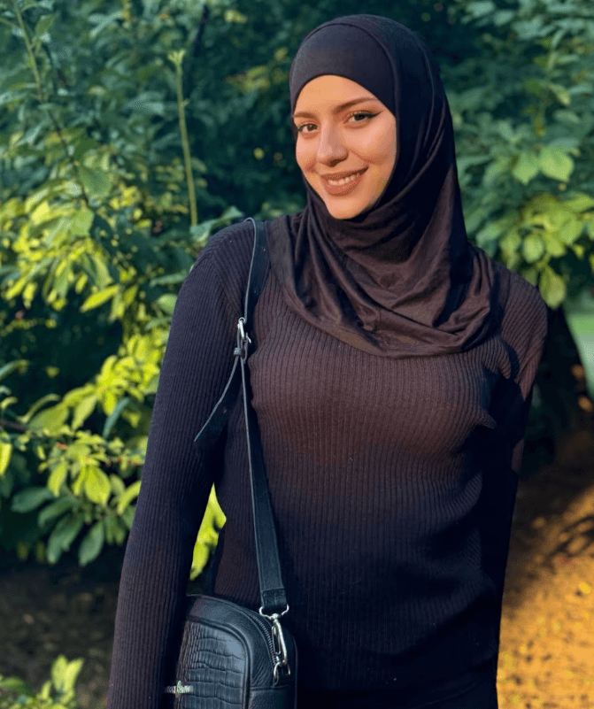 Smiling woman in a black hijab and ribbed sweater, standing among green foliage in a park with a small black crossbody bag.