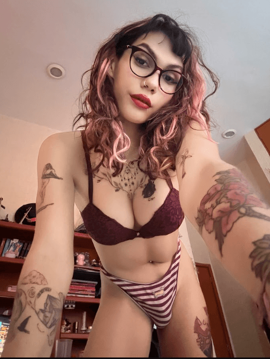 Person with pink-dyed hair, glasses, burgundy bra and striped panties posing for a room selfie.